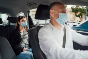 masked taxi driver and female passenger wearing seatbelt inside moving vehicle