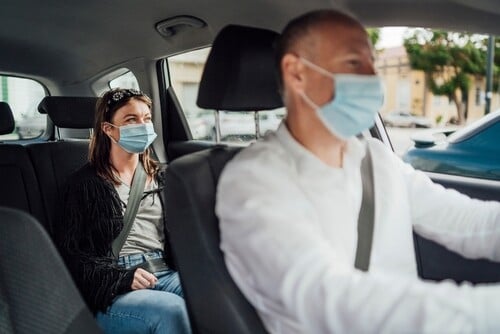 masked taxi driver and female passenger wearing seatbelt inside moving vehicle