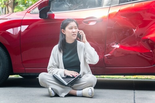 woman sitting on ground calling phone beside dented damaged car